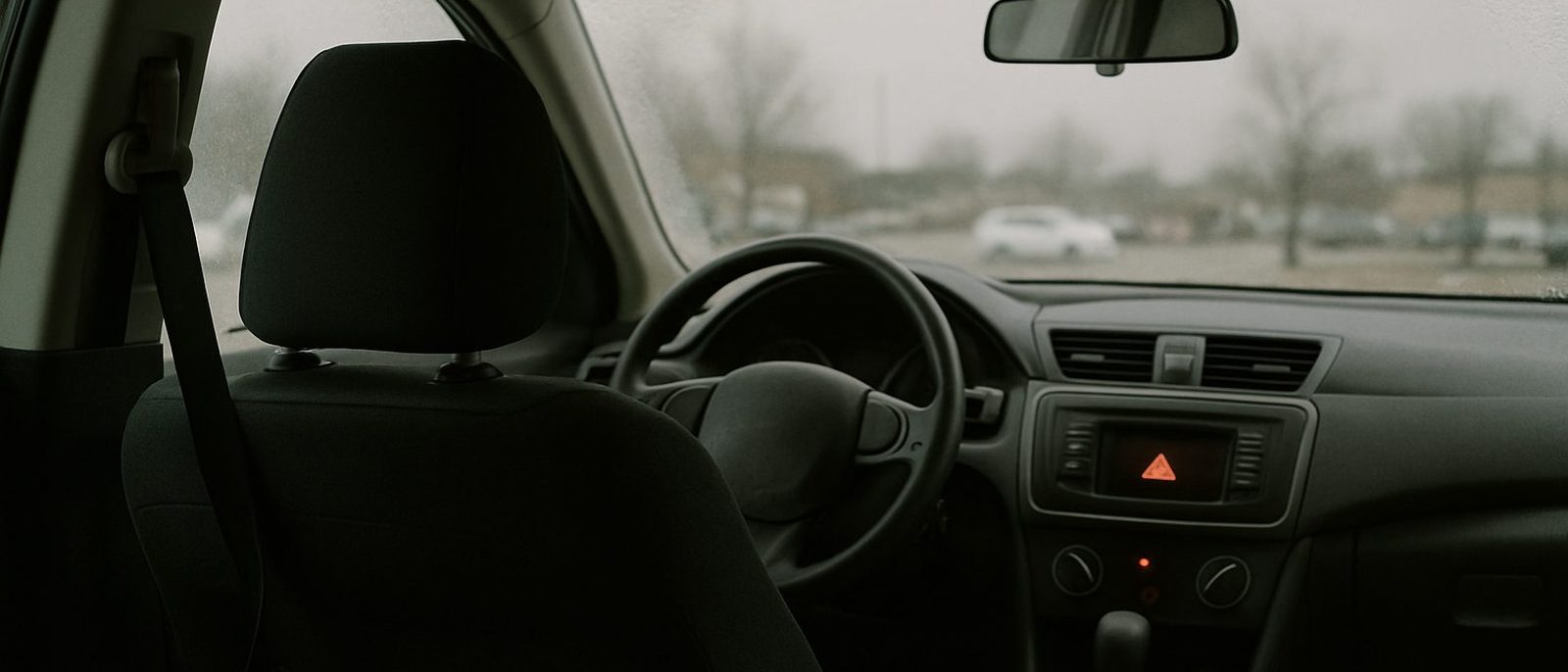 The interior of a parked sedan seen from the back seat, seat belt slack, hazard light glowing on the dash, grey parking lot beyond